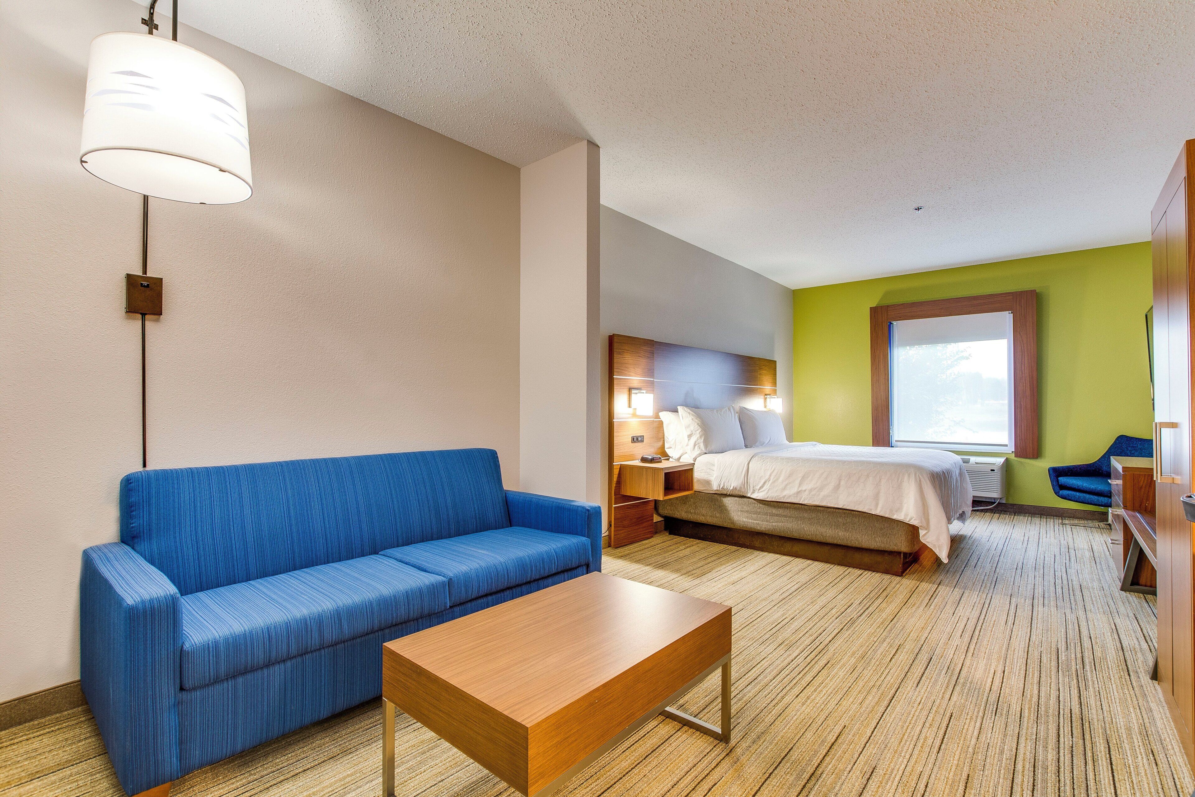 Una habitación de hotel con un sofá azul, una mesa de centro de madera, una cama con cabecera de madera, una mesita de noche y una ventana con vistas.