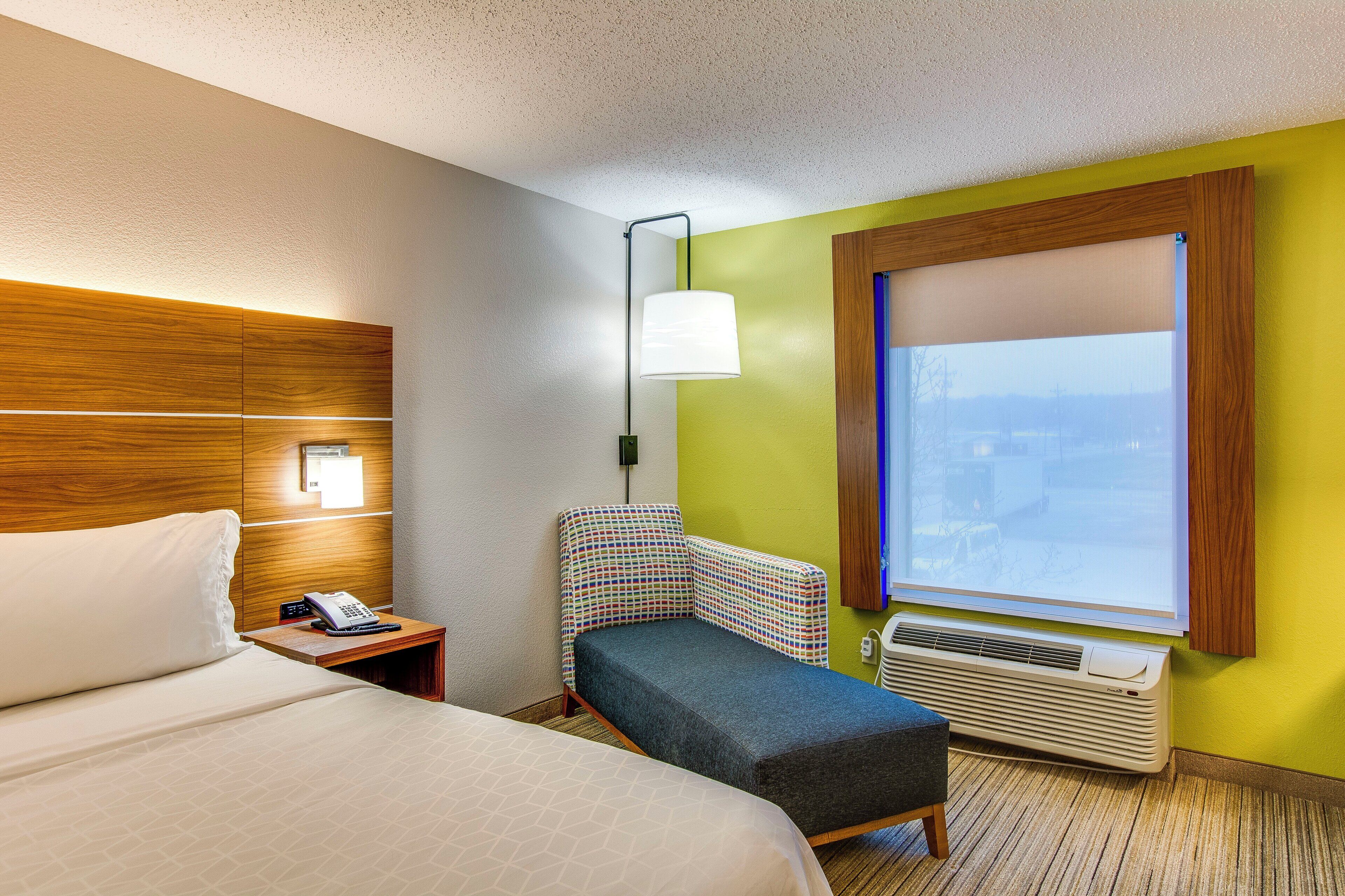 A hotel room with a bed, a desk with a telephone, a chair, a window with blinds, and an air conditioning unit.