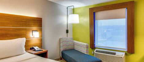 A hotel room with a bed, a desk with a telephone, a chair, a window with blinds, and an air conditioning unit.