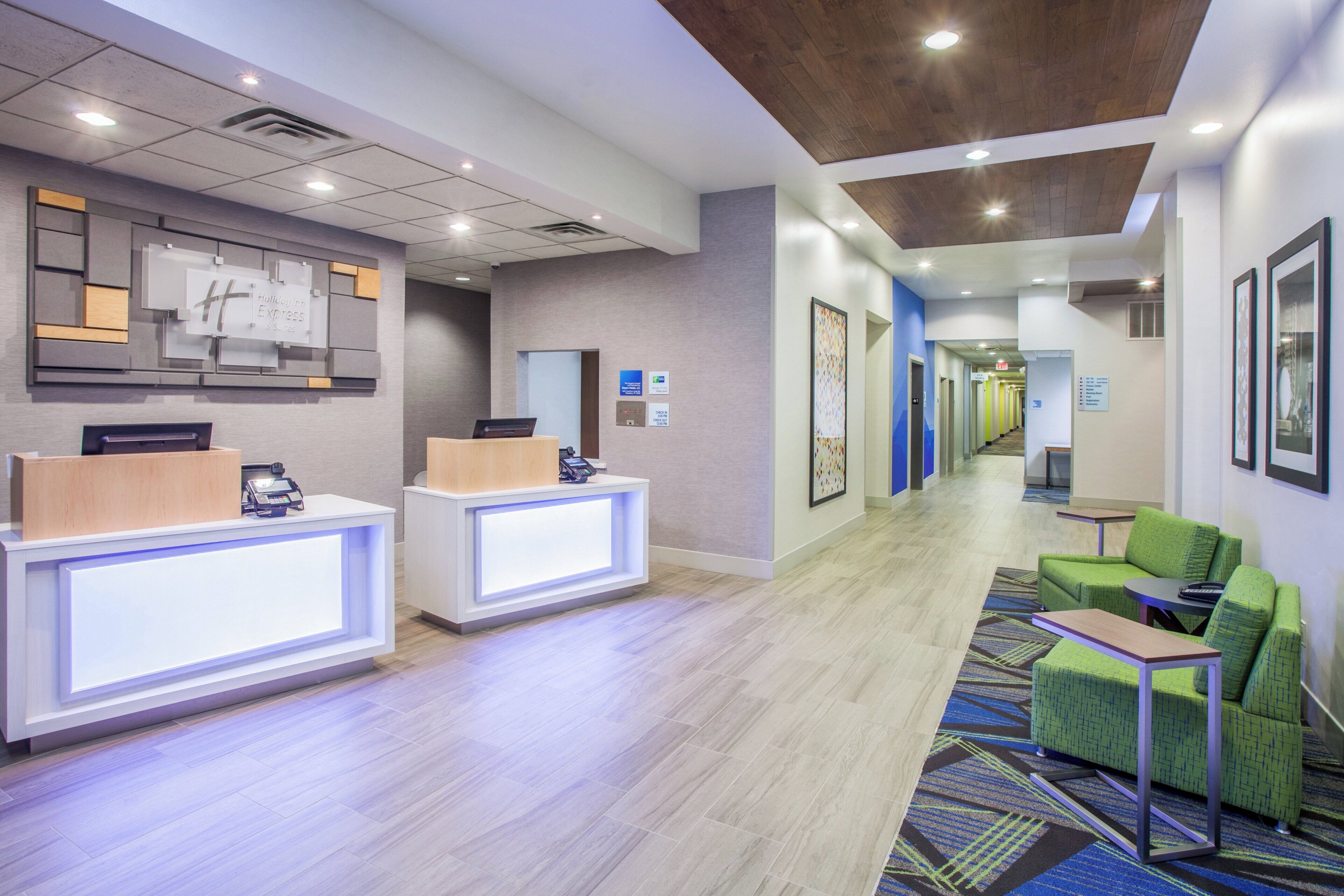 A hotel reception area with two reception desks, a waiting area with green seating, and a hallway with directional signs.
