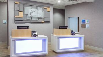 Zona de recepción de hotel con dos mostradores, un panel informativo en la pared y un quiosco de registro de entrada y salida.