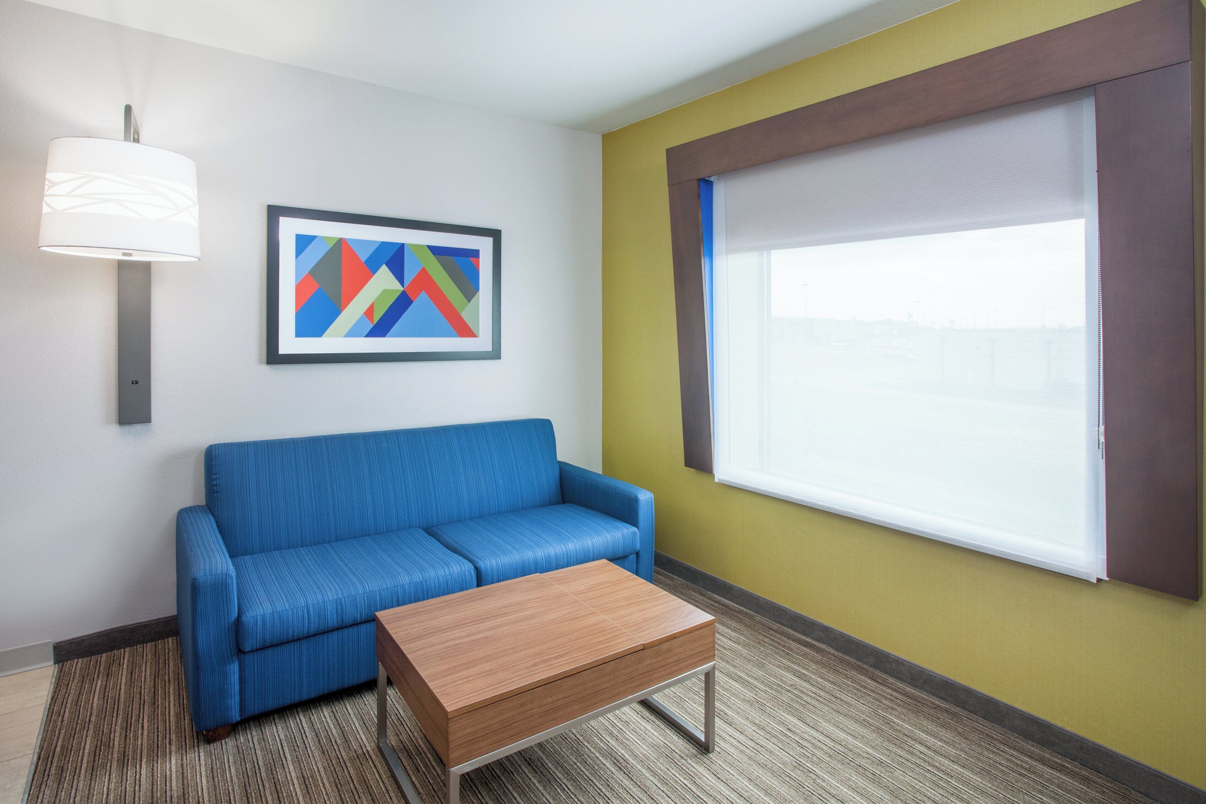 A hotel room with a blue sofa, a wooden table, a framed abstract painting, a wall-mounted lamp, and a window with blinds.