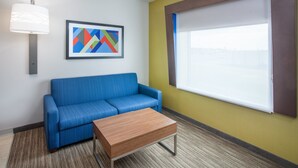 A room with a blue sofa, a wooden coffee table, a lamp, a framed abstract painting, and a window with blinds. - Holiday Inn Express & Suites Texarkana by IHG (Texarkana)