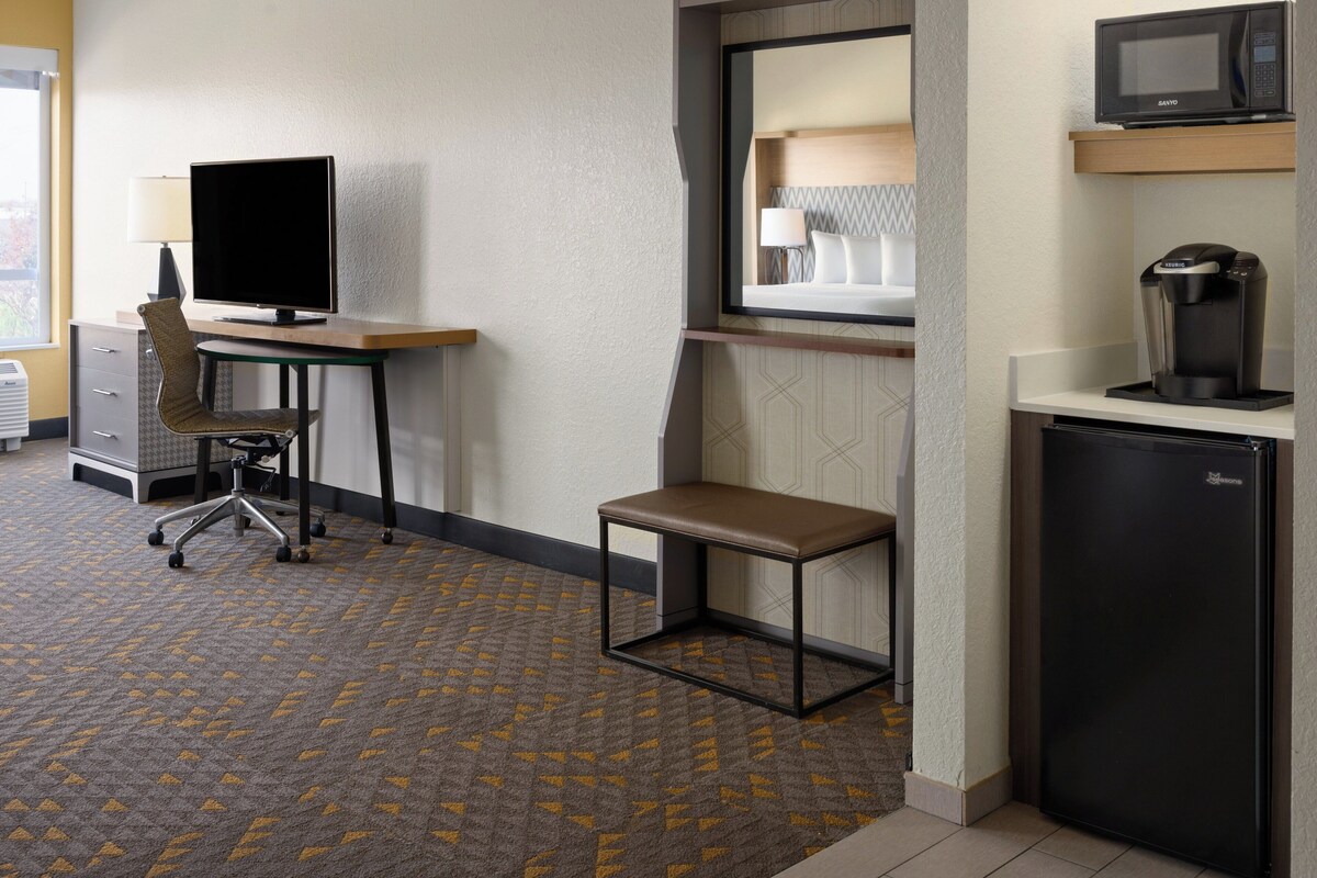 A hotel room with a desk, chair, and a small table. There is a microwave and a refrigerator.