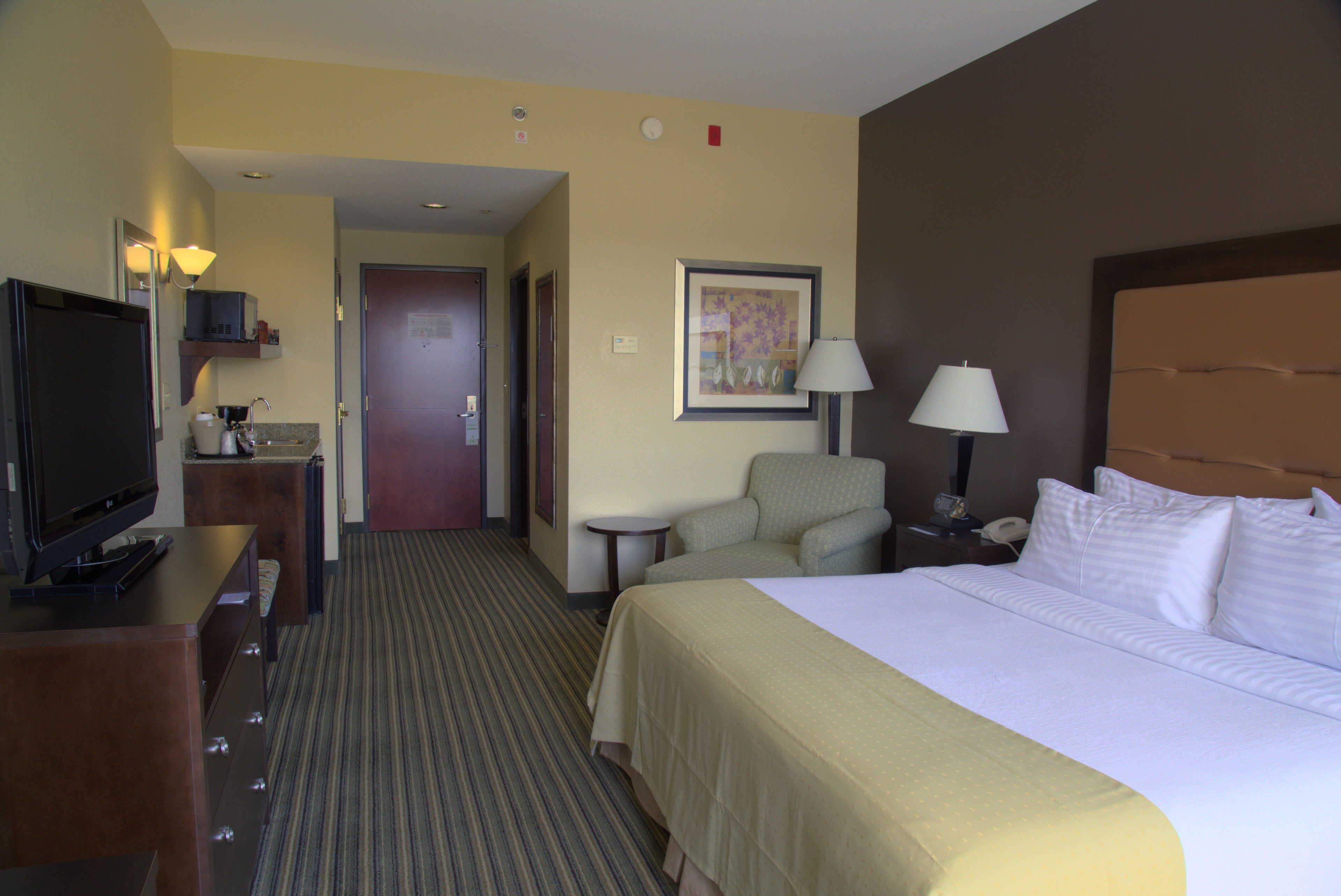 A hotel room with a bed, a TV, a desk with a lamp, a chair, and a painting on the wall.