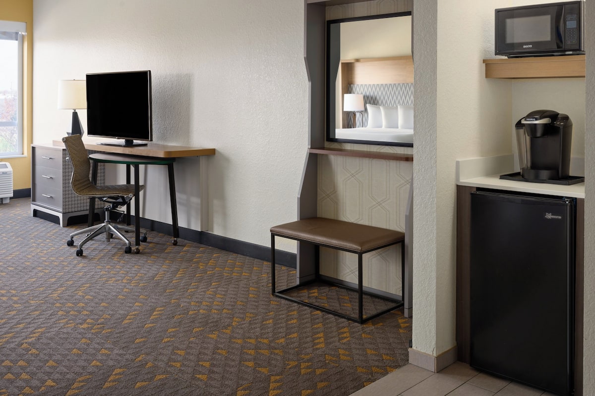 A hotel room with a desk, chair, and a small table. There is a microwave and a refrigerator.