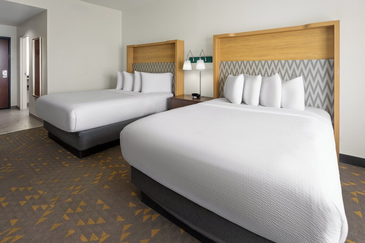 A hotel room with two beds, a wooden headboard, a bedside table with a lamp, and a patterned carpet.