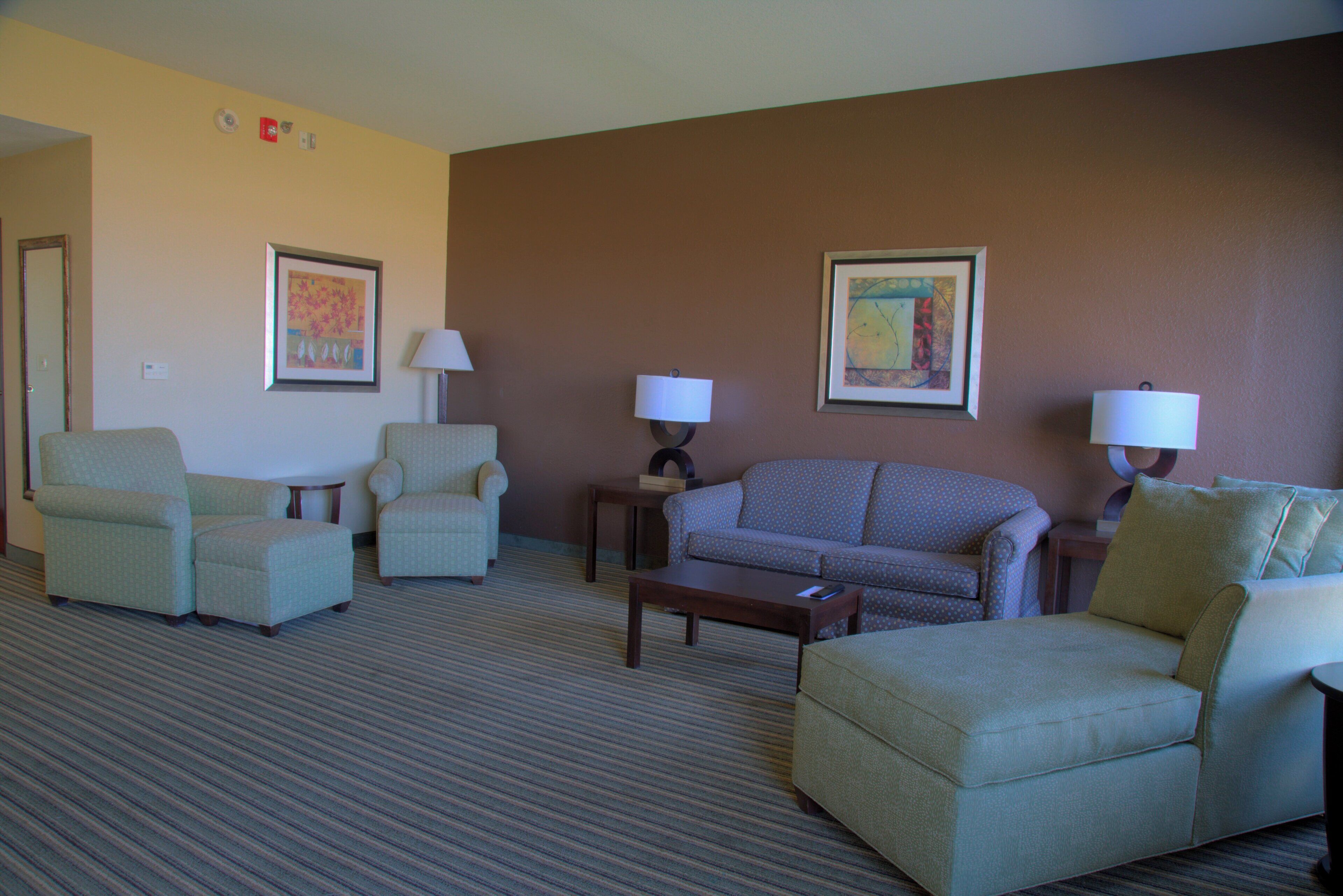 A hotel room with a seating area including two armchairs, a sofa, and a coffee table. There are two wall-mounted lamps and two framed pictures. The room has a carpeted floor and a door on the left.