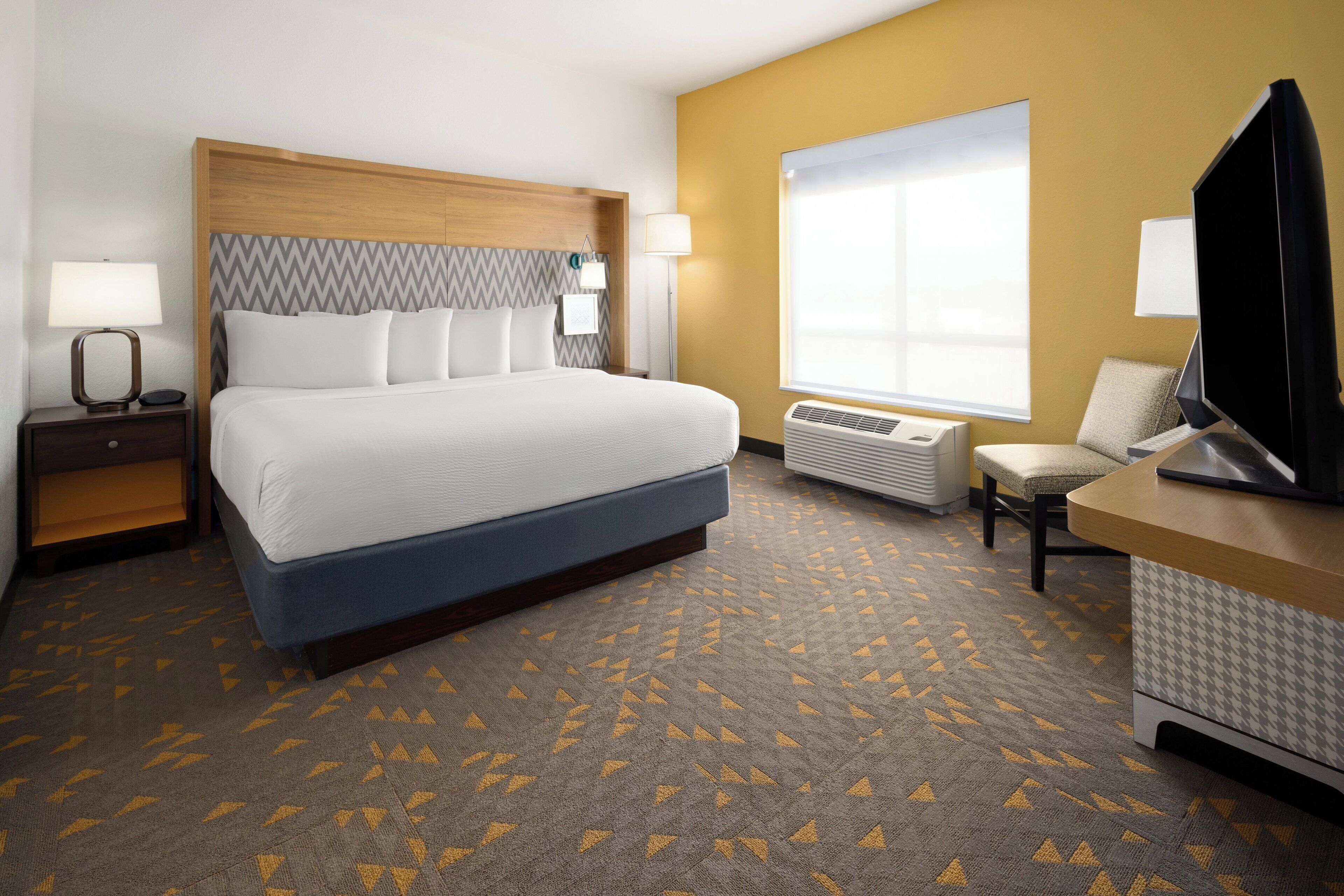 A hotel room with a large bed, a desk with a flat-screen TV, a chair, a nightstand with a lamp, and a window with blinds.
