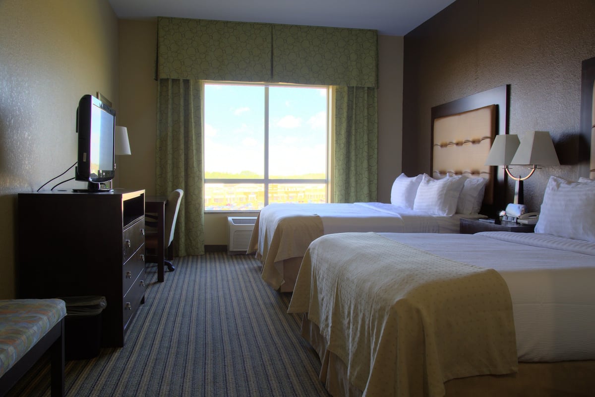 A hotel room with two beds, a desk with a computer, a window with curtains, and a nightstand with a lamp.