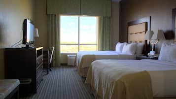 A hotel room with two beds, a desk with a computer, a window with curtains, and a nightstand with a lamp.