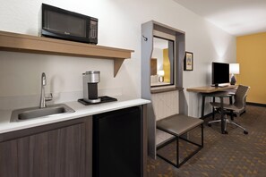 A compact hotel room with a kitchenette, including a microwave, coffee maker, and sink. There is a desk with a computer and chair, and a small bench.
