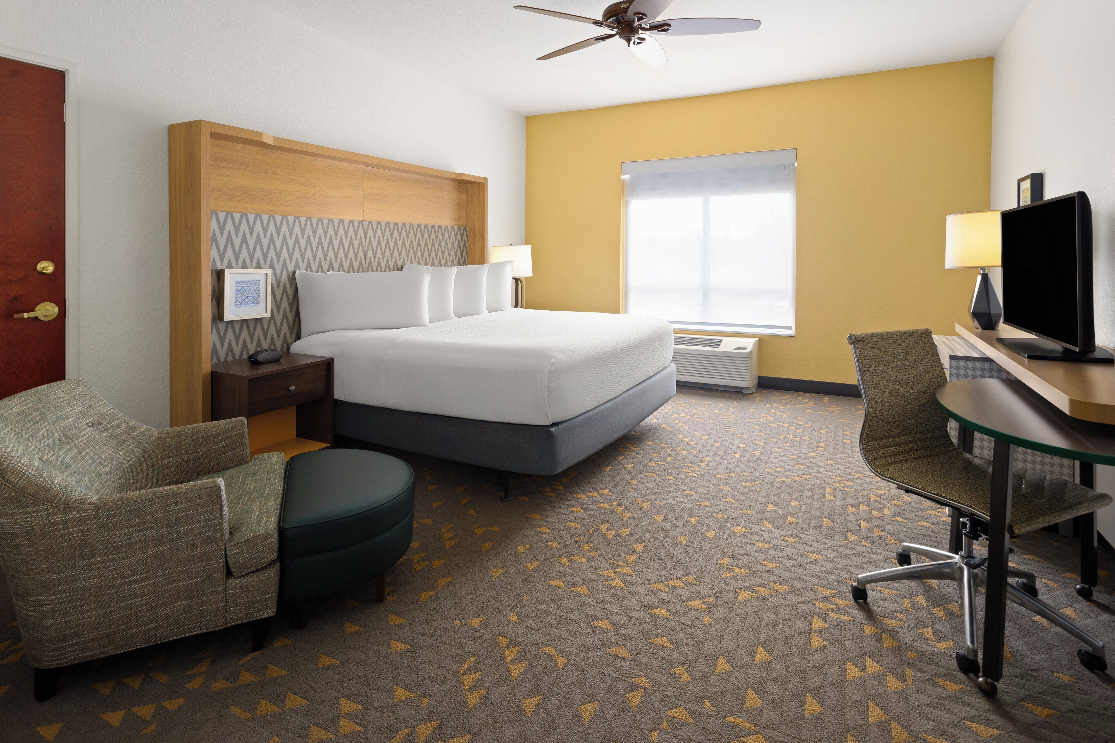 Une chambre d’hôtel équipée d’un lit, d’un bureau avec un ordinateur, d’une chaise, d’une télévision, d’un ventilateur de plafond et d’une fenêtre avec des stores.
