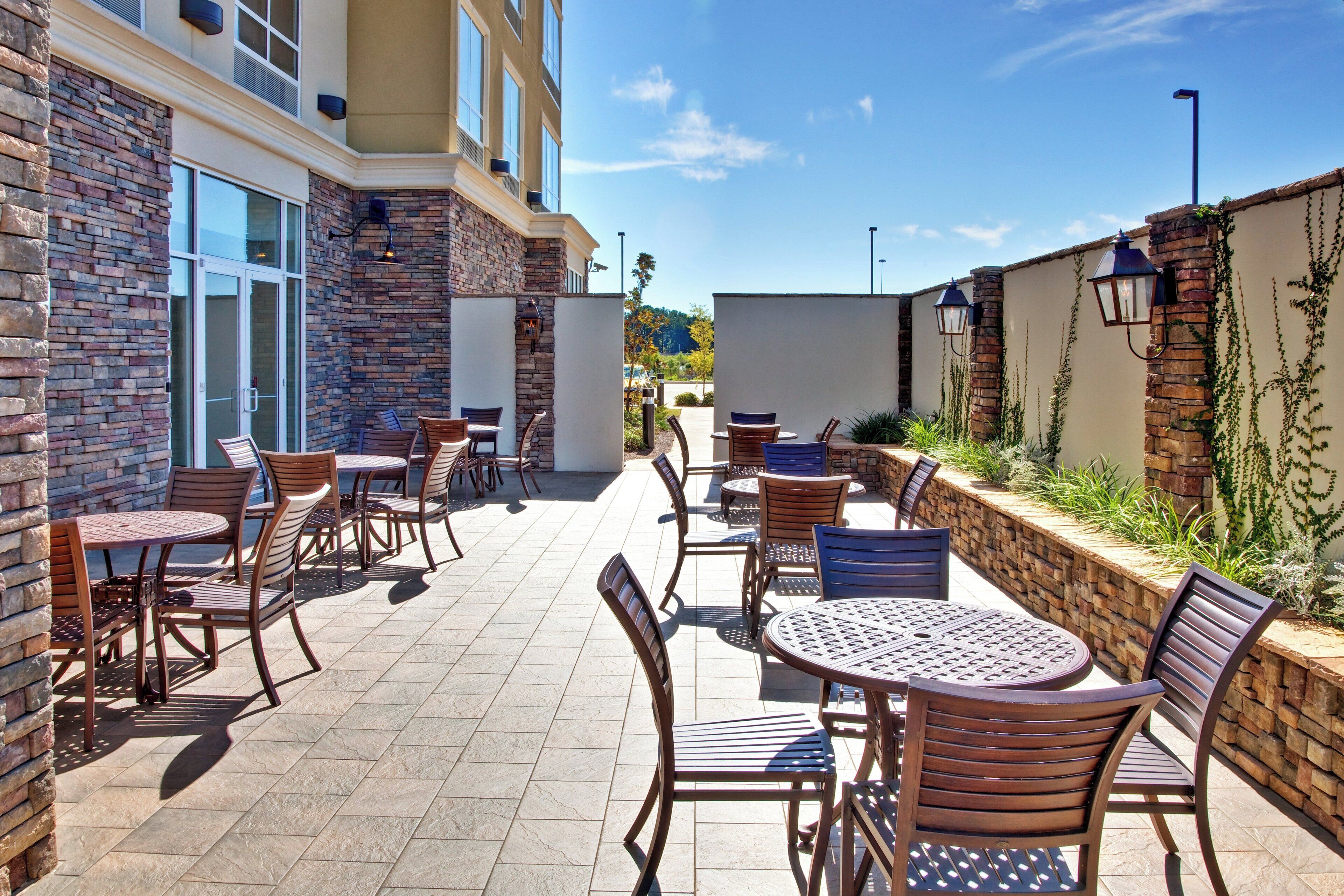 Un espace extérieur aménagé en terrasse, avec plusieurs tables et chaises, un mur en pierre et un bâtiment doté de grandes fenêtres.