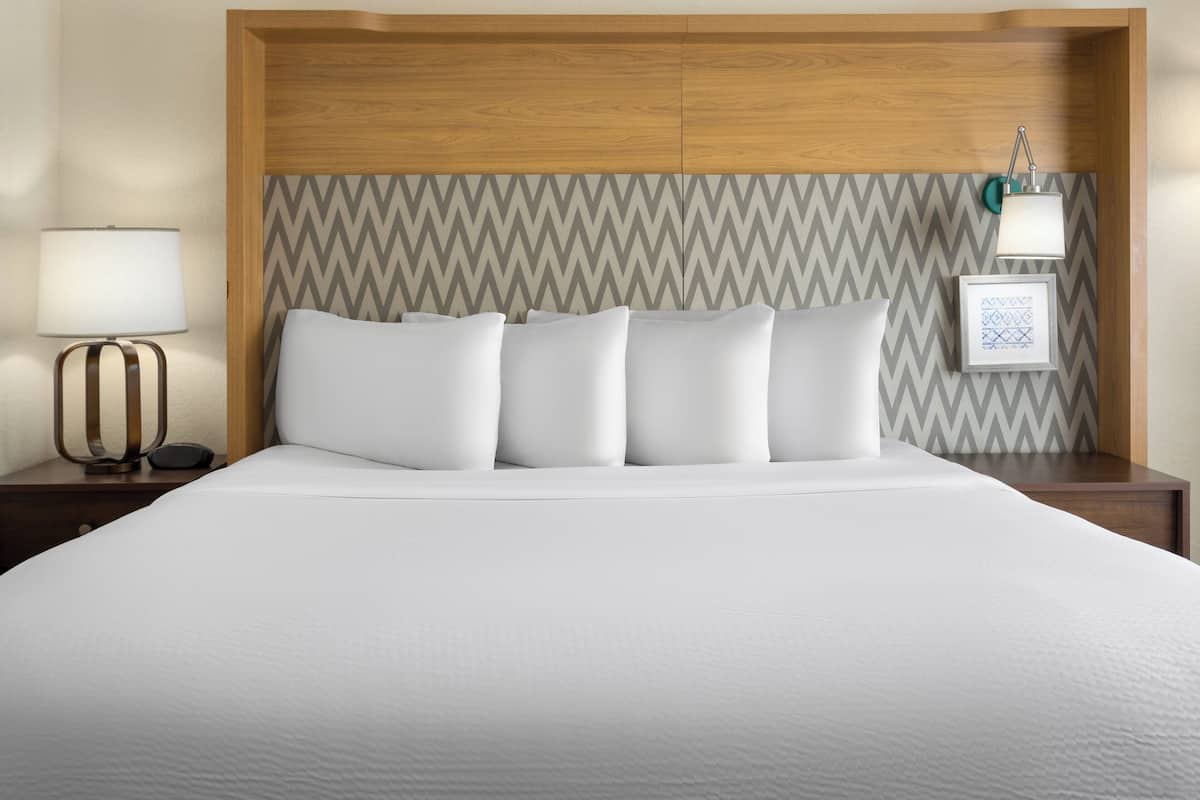 A neatly made bed with white linens and pillows, a wooden headboard, a bedside lamp, and a framed picture on the wall.