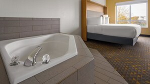 A modern hotel room with a large bathtub, a bed with a wooden headboard, and a view of the outdoors through a large window. - Holiday Inn Jackson Southeast - Pearl by IHG (Pearl)