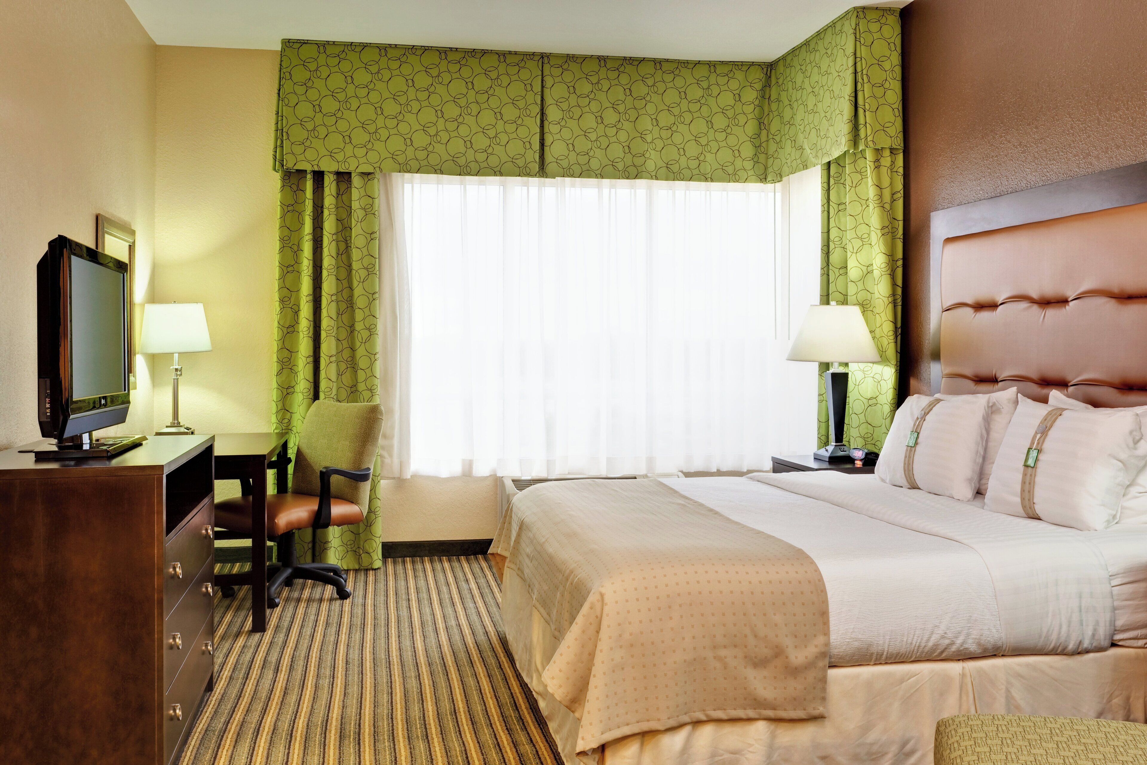 A hotel room with a bed, a desk with a television, two bedside lamps, and a window with curtains.