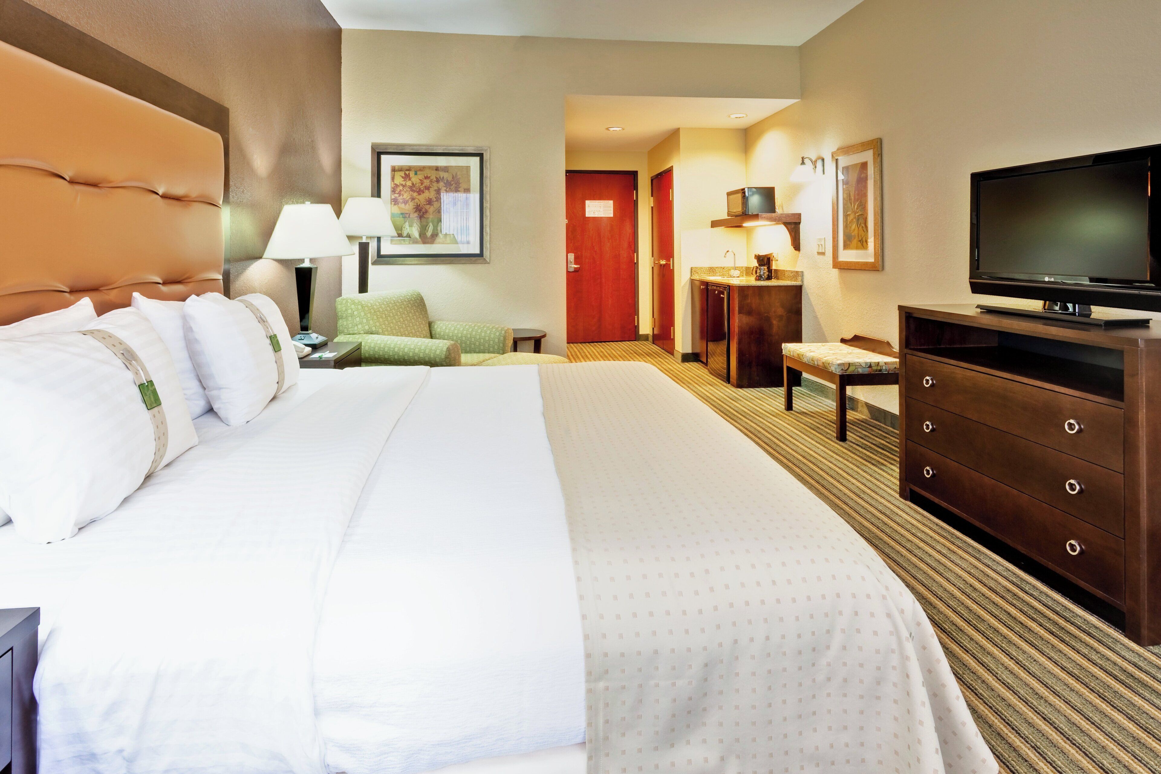 A hotel room with a large bed, a television, a desk with a chair, and a small kitchenette.