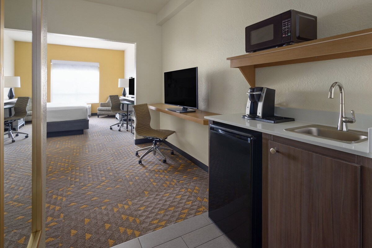 A hotel room with a kitchenette, a bed, a desk, a chair, and a television.