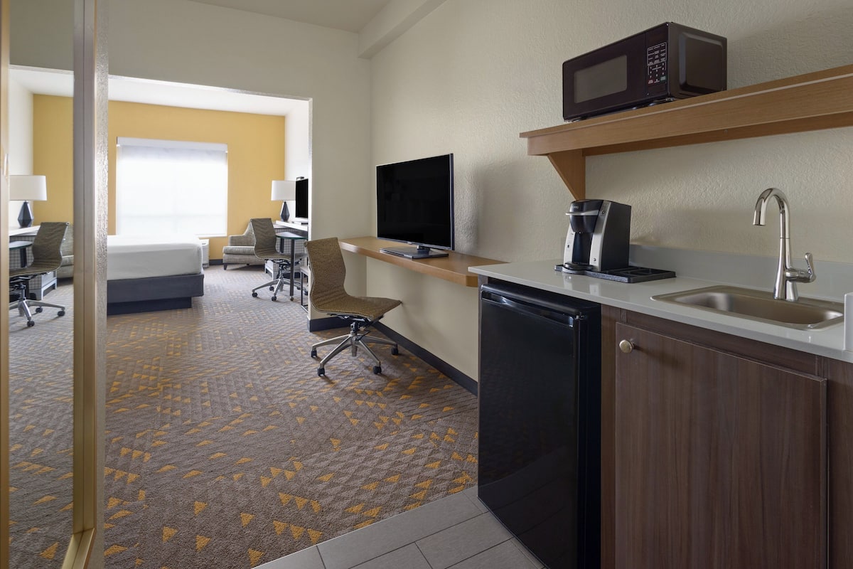 A hotel room with a kitchenette, a bed, a desk, a chair, and a television.