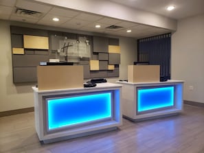 A reception desk with a blue illuminated panel, a wall-mounted information board, and a window with blinds. - Holiday Inn Express & Suites Lincoln Airport by IHG (Lincoln)