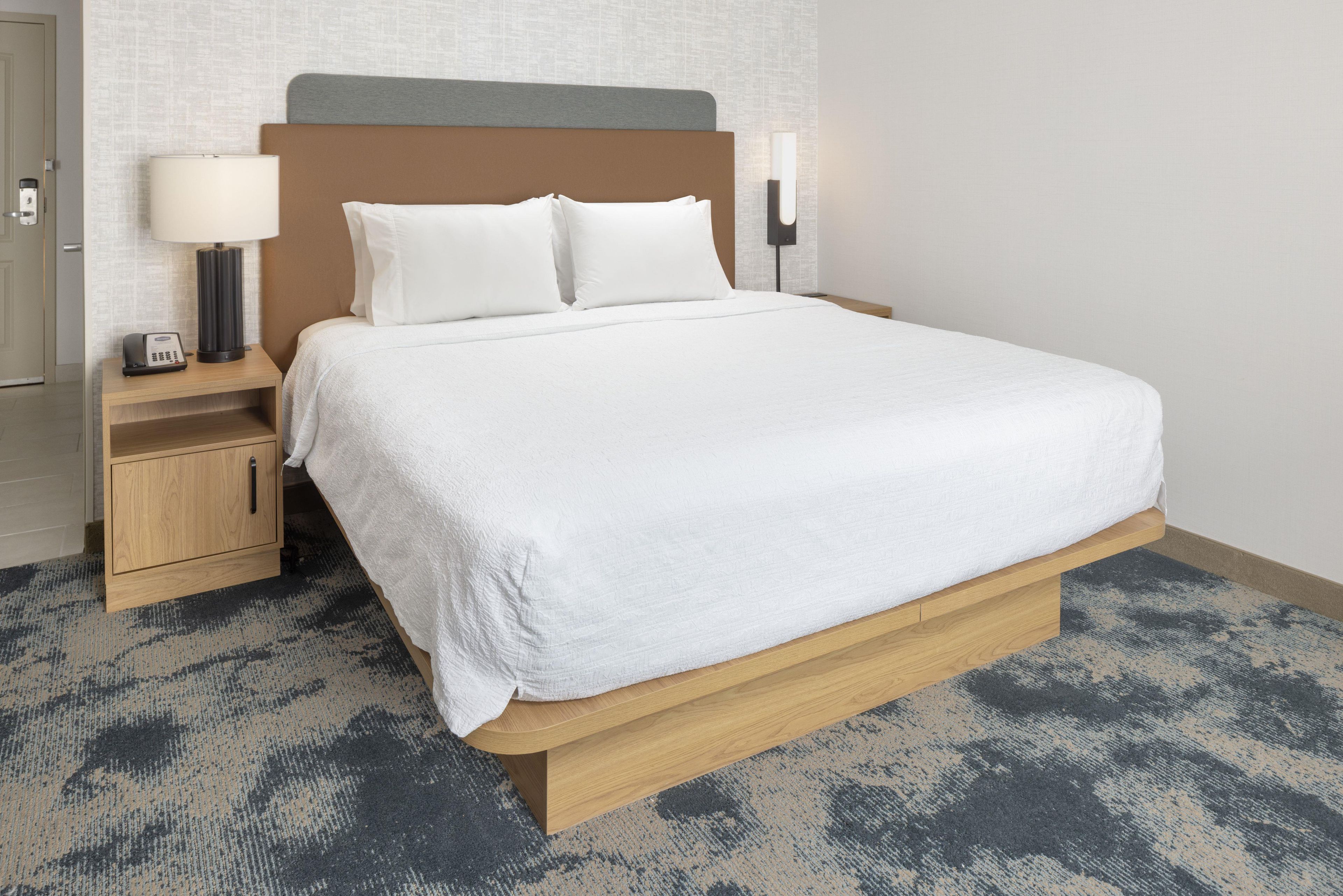 A hotel room with a bed, nightstand, lamp, and wall-mounted light.