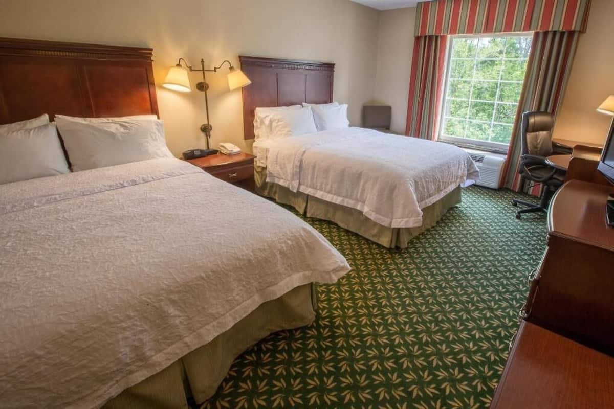 A hotel room with two beds, a desk, a chair, a lamp, and a window with curtains.