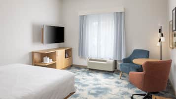 A hotel room with a bed, a desk, a chair, a television, a window with curtains, and an air conditioning unit.