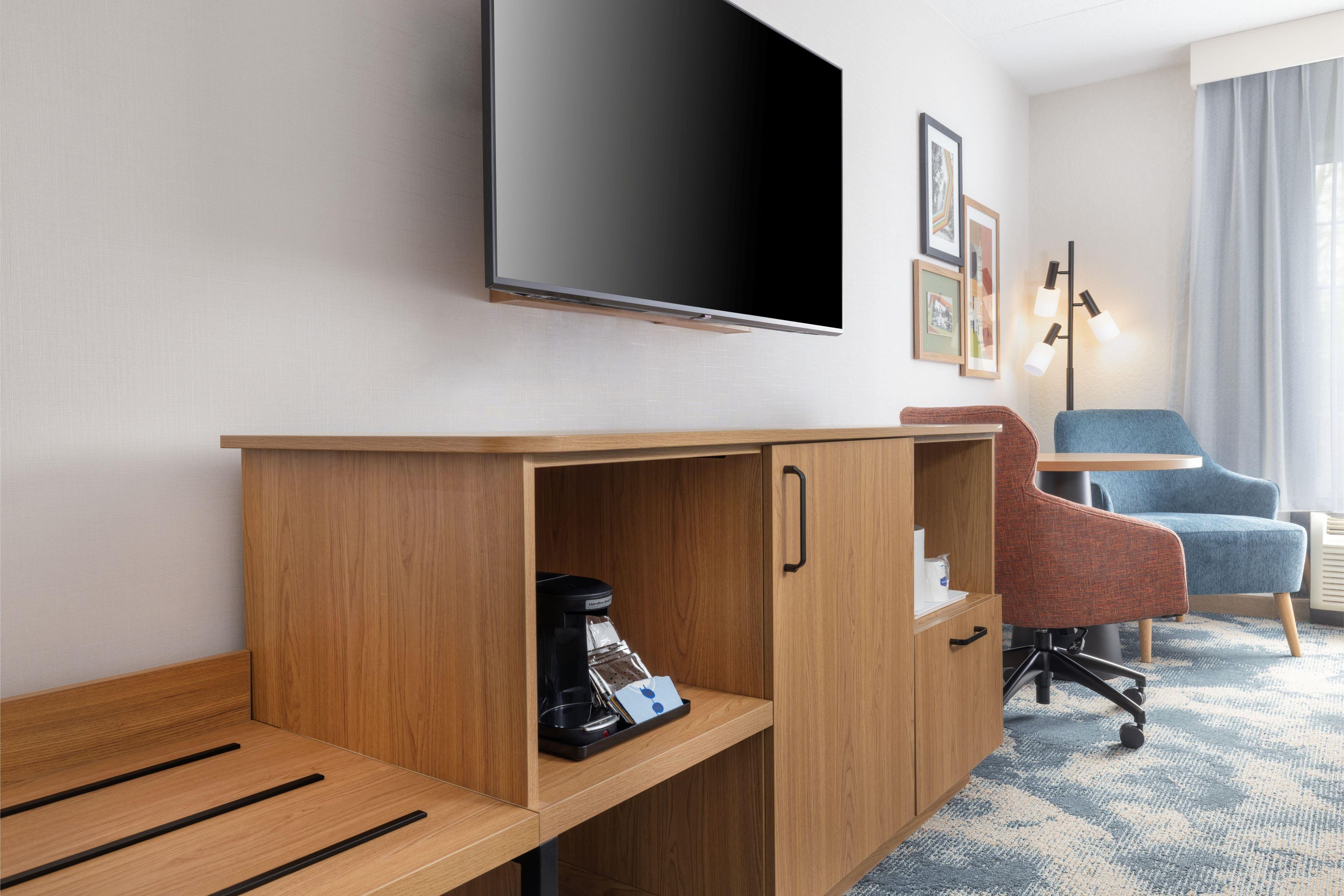 A hotel room with a wooden desk, a flat-screen TV, a coffee maker, a blue chair, and a lamp.