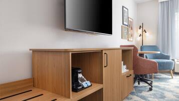 A hotel room with a wooden desk, a flat-screen TV, a coffee maker, a blue chair, and a lamp.