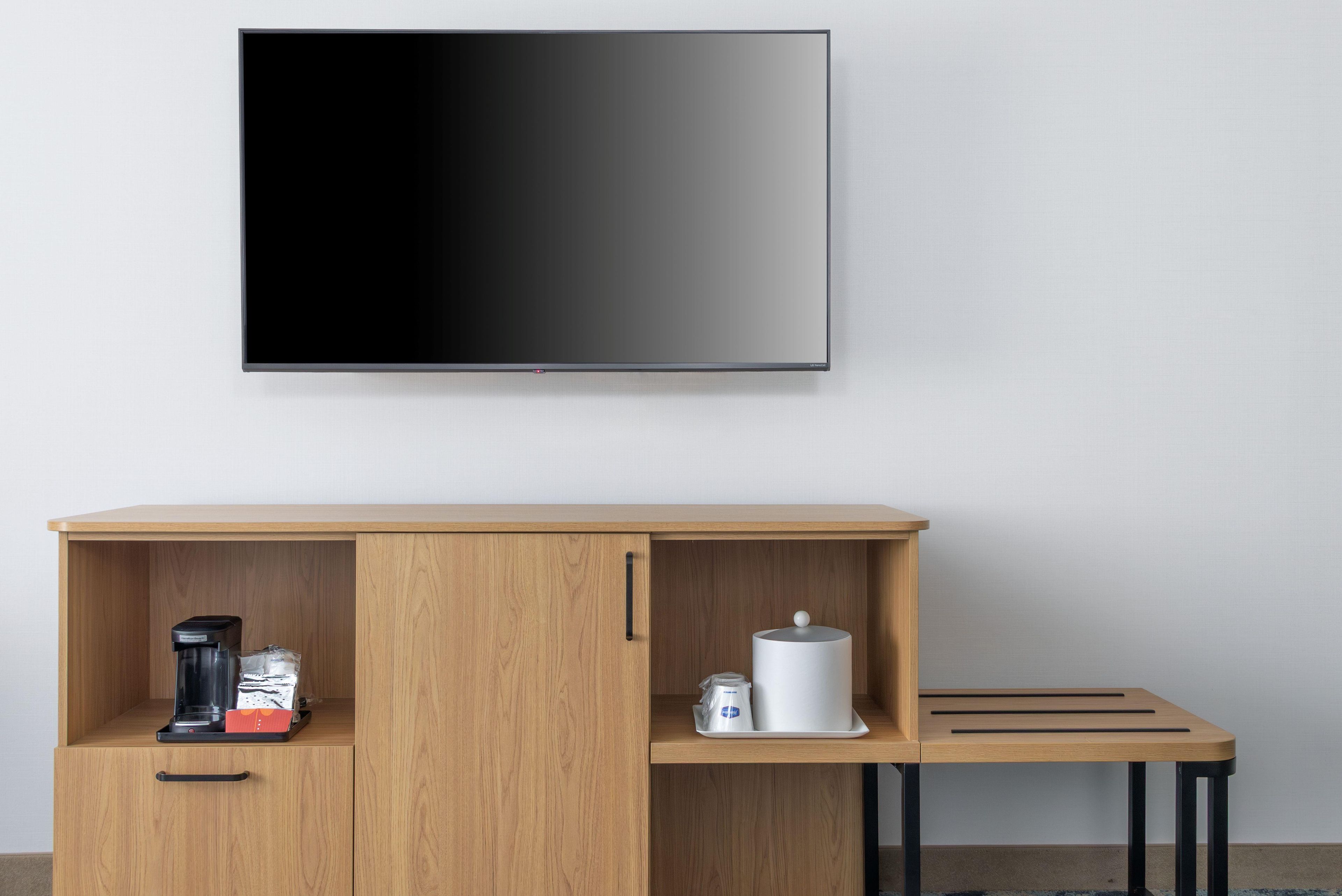 A room with a flat-screen TV mounted on the wall, a wooden cabinet with a coffee maker, a kettle, and a white container.