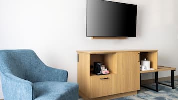 A room with a blue armchair, a wooden cabinet with a coffee maker, a television on the wall, and a small table with a cup.