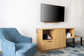 Ein Zimmer mit blauem Sessel, einer Holzkommode mit Kaffeemaschine, einem Fernseher an der Wand und einem kleinen Tisch mit einer Tasse.