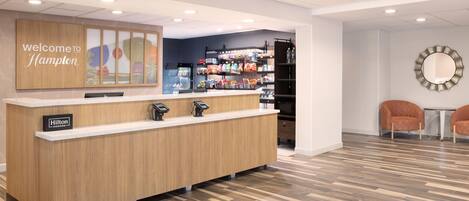 A hotel reception area with a wooden counter, a 'Welcome to Hampton' sign, a minibar, and two orange chairs.