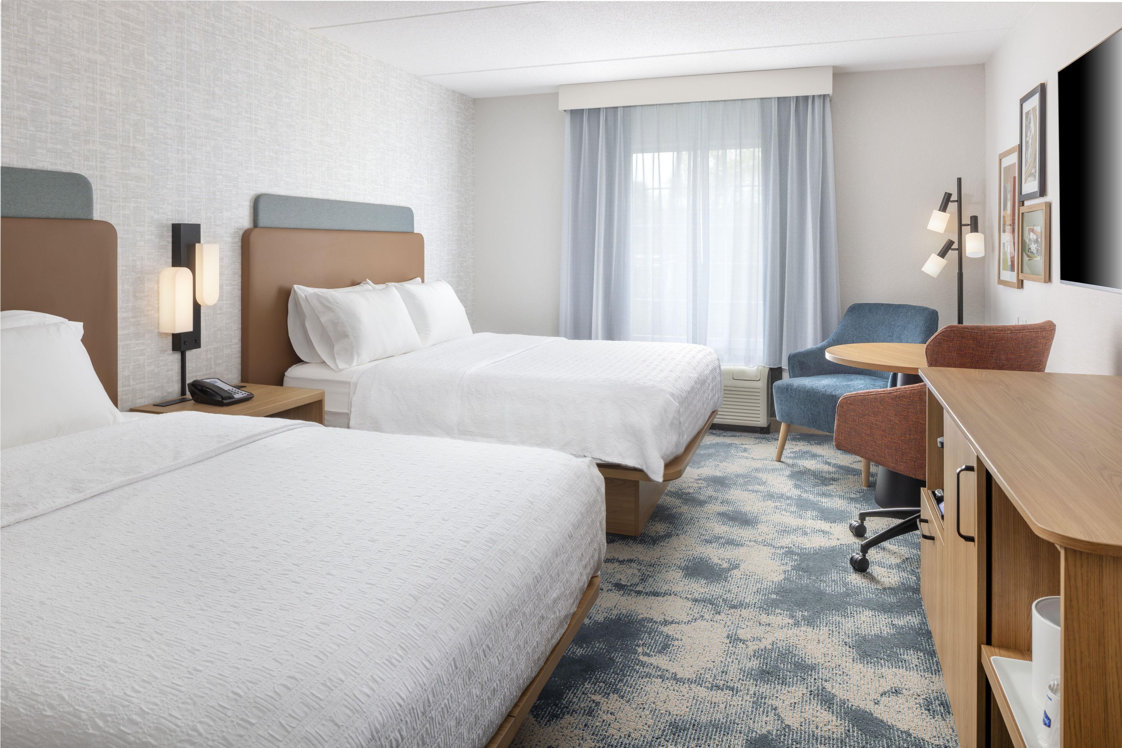 A hotel room with two beds, a desk, a chair, a lamp, and a large window with curtains.