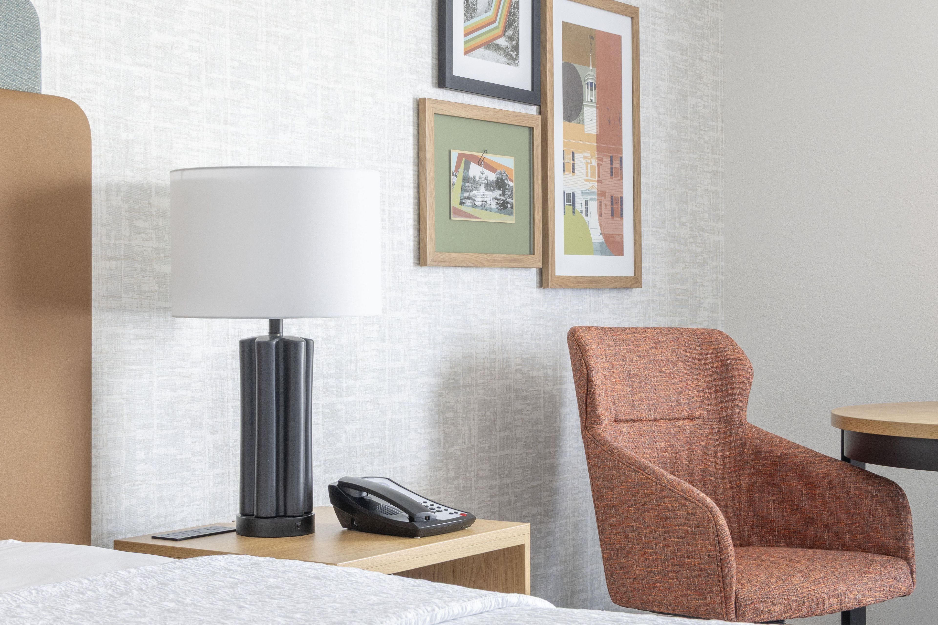A hotel room with a bed, a nightstand with a lamp and phone, a chair, and framed pictures on the wall.