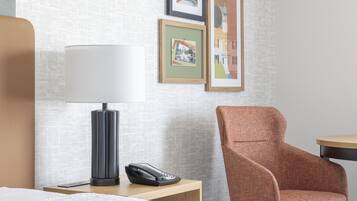 A hotel room with a bed, a nightstand with a lamp and phone, a chair, and framed pictures on the wall.