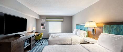 A hotel room with two beds, a flat-screen TV, a desk with a chair, a window, and a bathroom with a mirror and towel rack.