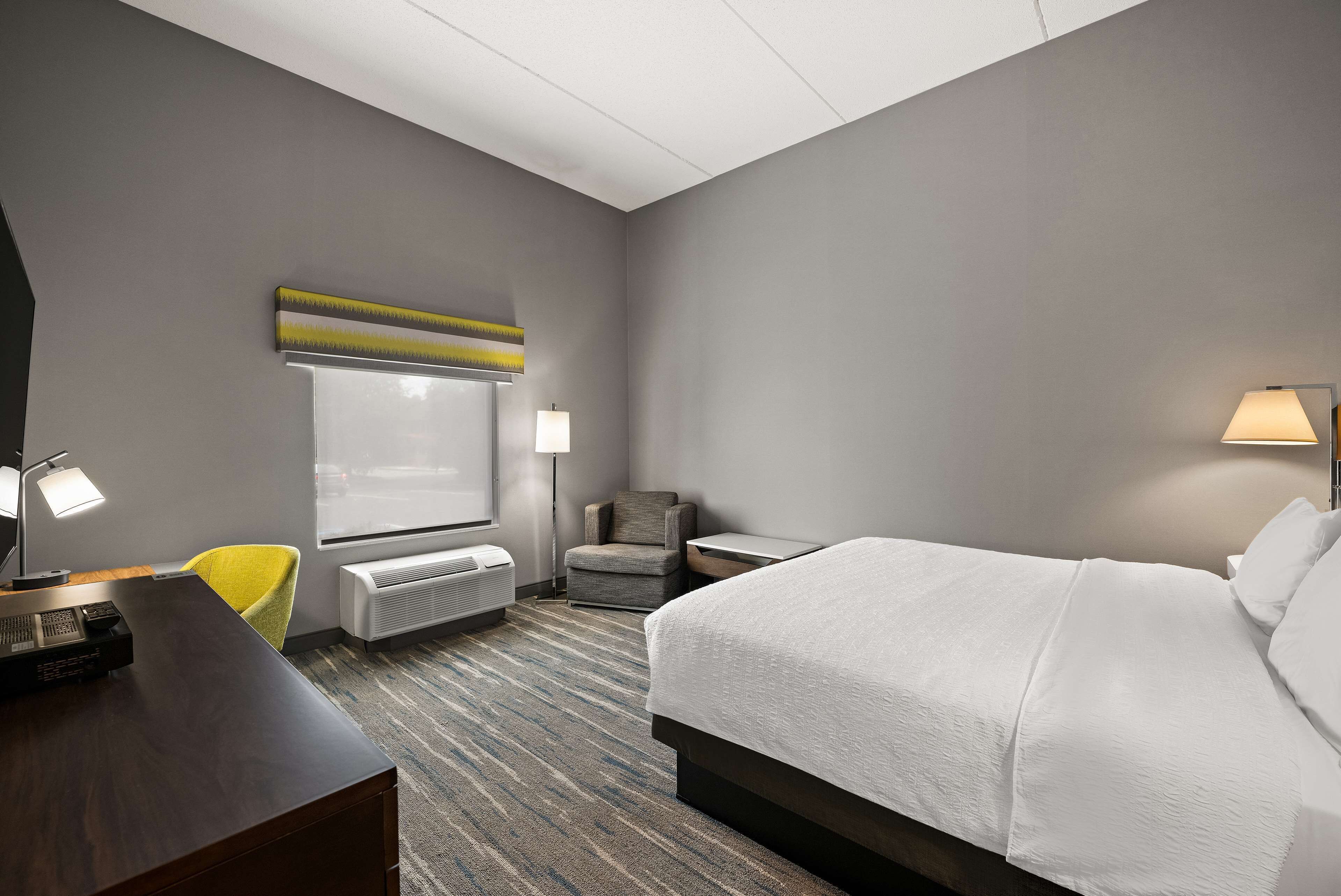 A hotel room with a large bed, a desk with a computer, a chair, a lamp, and a window with blinds.