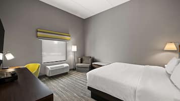 A hotel room with a large bed, a desk with a computer, a chair, a lamp, and a window with blinds.