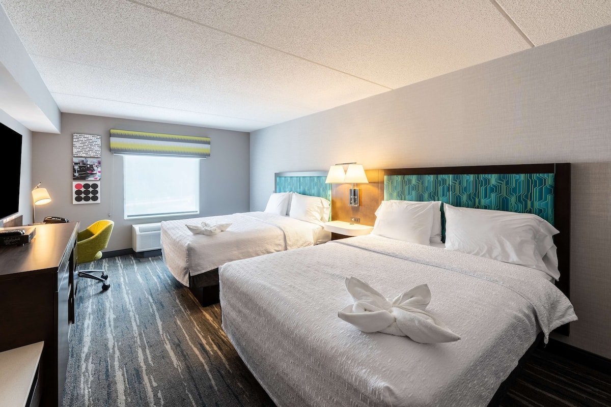A hotel room with two beds, a desk, a chair, and a window.