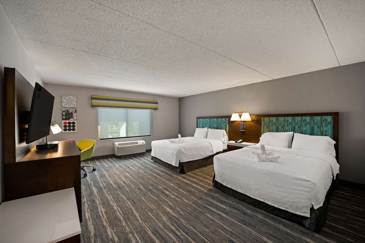 A hotel room with two beds, a desk with a computer, a TV, a window, and a wall-mounted heater.