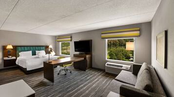Ein Hotelzimmer mit Bett, Schreibtisch, Fernseher, Sofa und großen Fenstern mit Blick auf Grünflächen.