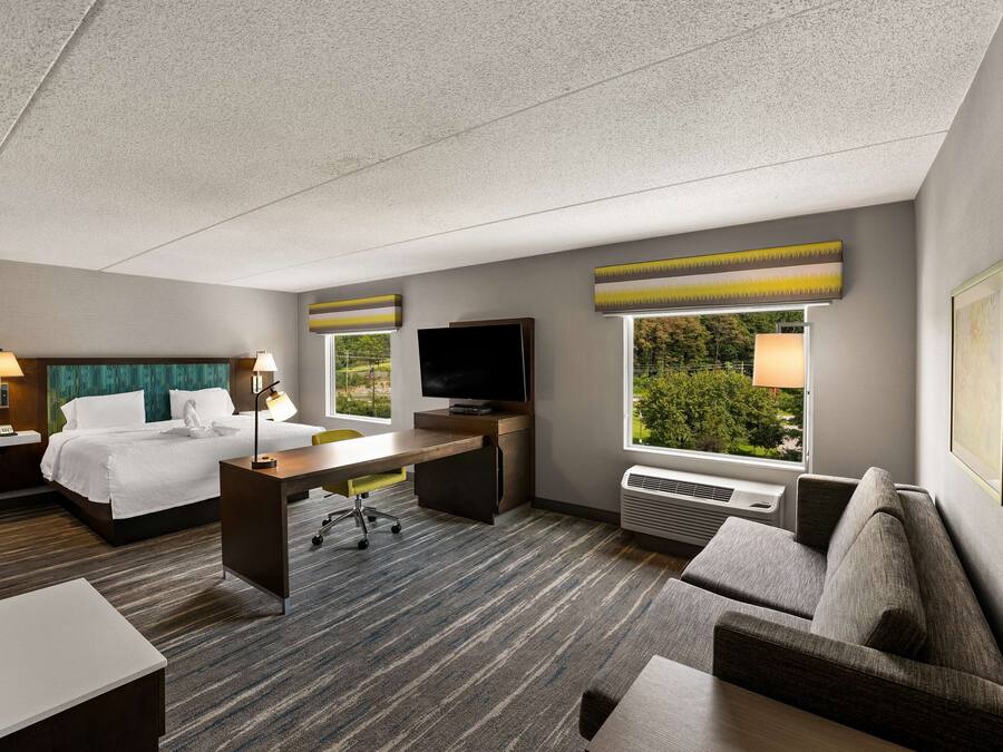 Ein Hotelzimmer mit Bett, Schreibtisch, Fernseher, Sofa und großen Fenstern mit Blick auf Grünflächen.