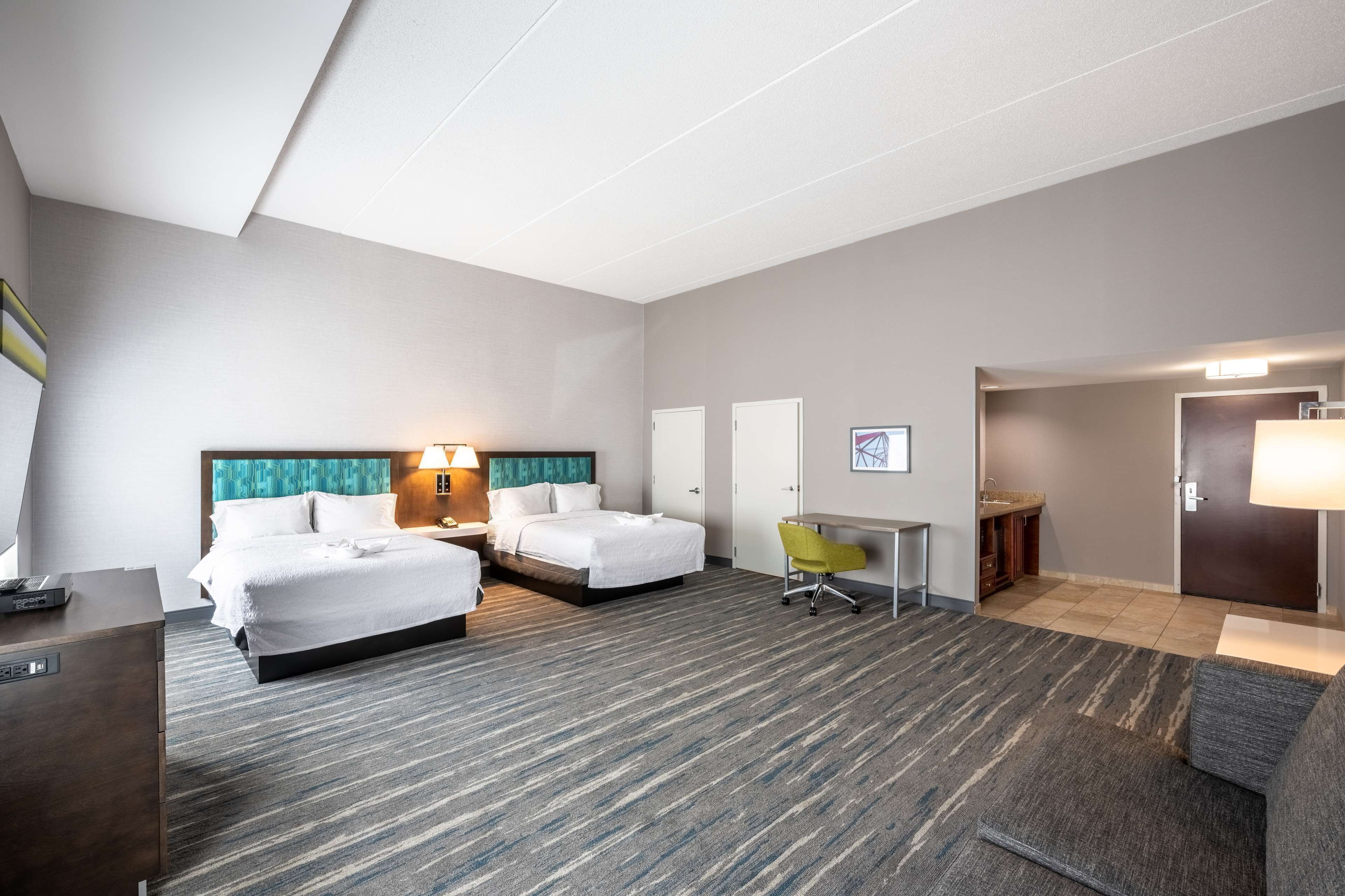A hotel room with two beds, a desk, a chair, and a small kitchenette.