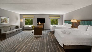 A hotel room with a large bed, a sofa, a desk, and a TV. There are windows with curtains, a lamp, and a picture on the wall.