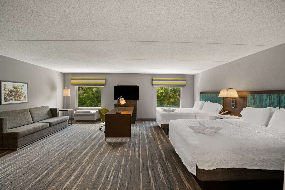 A hotel room with a large bed, a sofa, a desk, and a TV. There are windows with curtains, a lamp, and a picture on the wall.