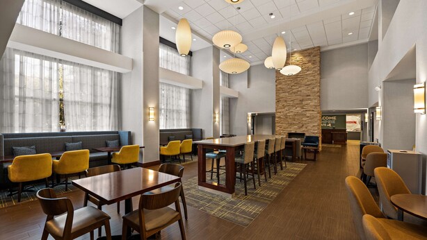 A modern hotel lobby with wooden flooring, yellow chairs, a long dining table, and pendant lighting.