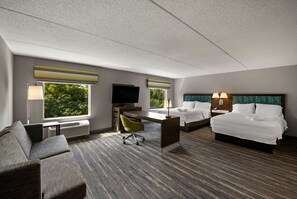 A hotel room with a sofa, a desk, a TV, two beds, and large windows with a view of trees.