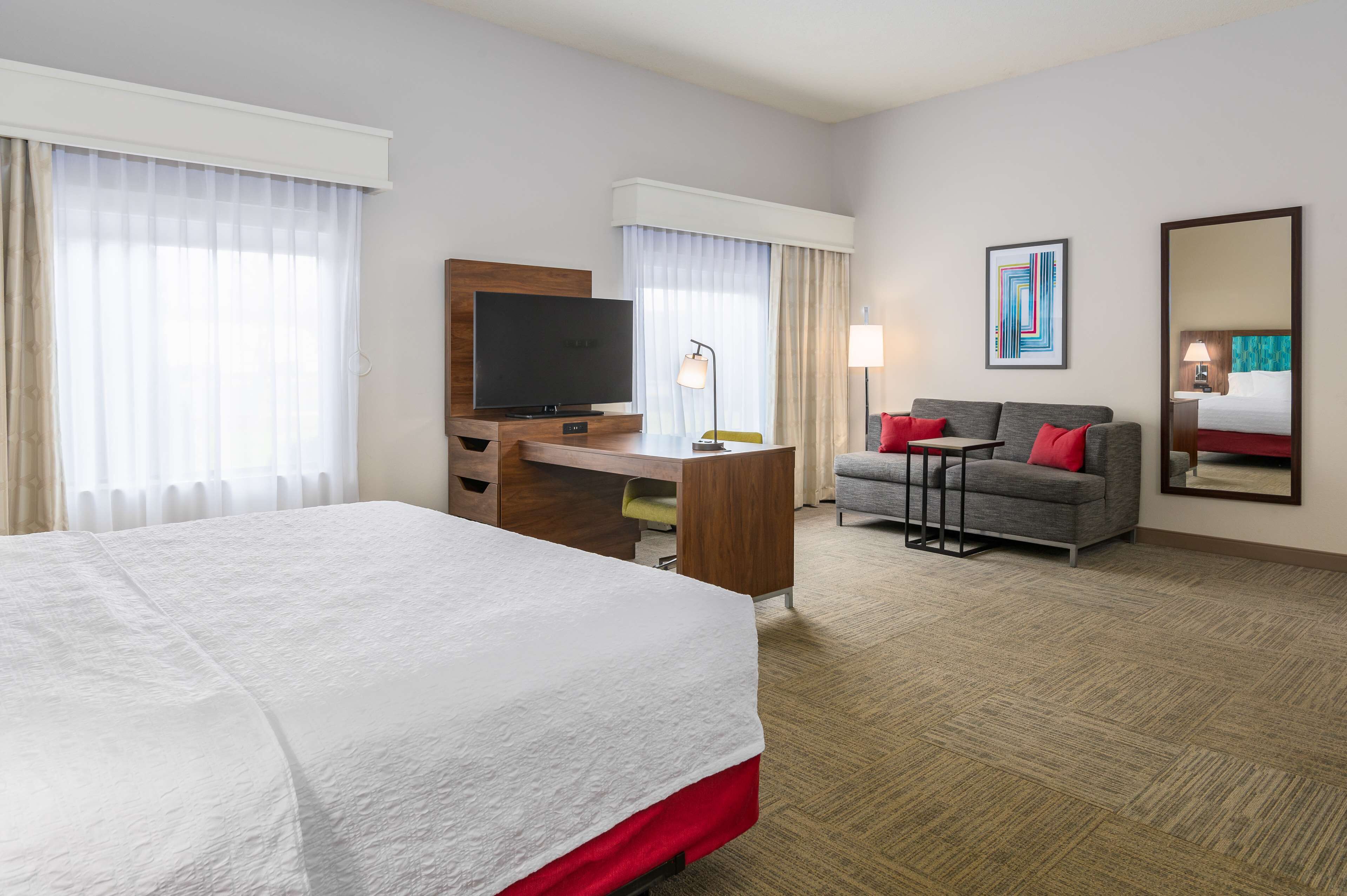A hotel room with a large bed, a desk with a TV, a sofa with red cushions, and a large mirror.